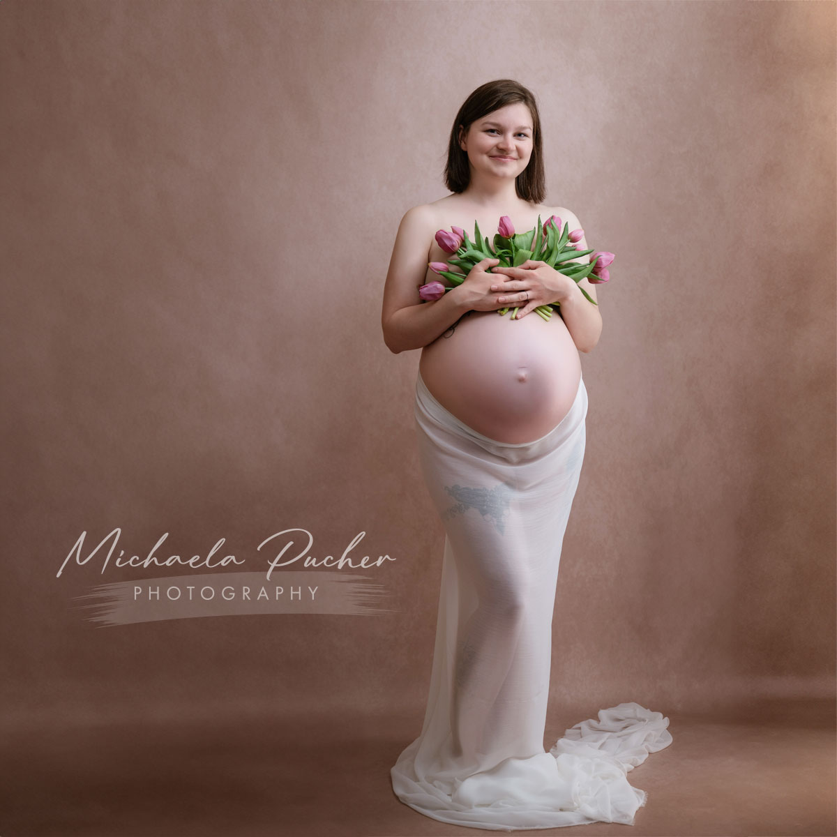 Schwangere Frau steht vor einem warmen, braunen Hintergrund, hält einen Strauß rosa Tulpen über ihrem nackten Babybauch und ist in ein zartes weißes Tuch gehüllt.